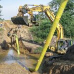 Ein gelber Bagger hebt ein mit gelbem Schutzmaterial umwickeltes Rohrstück über einen Wassergraben. Mehrere Bauarbeiter stehen auf einer erhöhten Böschung und beobachten die Arbeiten.