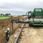 Ein grünes Baustellenfahrzeug schüttet Sand auf eine verlegte Pipeline, während ein Bauarbeiter in einer Grube mit einer Schaufel arbeitet. Im Hintergrund sind mehrere Personen zu sehen.