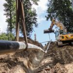 Eine Rohrleitung wird mit Gurten von einem Bagger in einen steilen Graben abgesenkt. Die Baustelle befindet sich in einem bewaldeten Hangbereich.