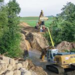 Ein gelber Bagger arbeitet in einem Graben mit Wasser, während ein Rohrstück in das Wasser abgesenkt wird. Zwei Bauarbeiter stehen auf der gegenüberliegenden Böschung und überwachen die Arbeiten.