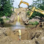 Zwei Bagger arbeiten an einer Flussquerung, bei der ein gelbes Rohrstück in den Graben abgesenkt wird. Ein Bauarbeiter steht auf der gegenüberliegenden Böschung.