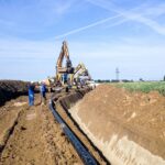 Zwei Bauarbeiter in blauen Overalls arbeiten an einer Pipeline-Verlegung in einem Erdgraben. Im Hintergrund befinden sich mehrere Bagger und eine Stromleitung.