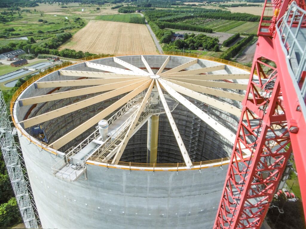 Ein Silo im Bau mit einer Fachwerkkonstruktion für das Dach. Der Kran hebt Baumaterial nach oben.