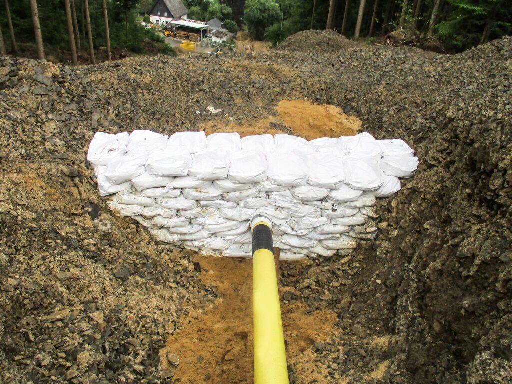 Eine Pipeline mit einer Barriere aus Sandsäcken, die wahrscheinlich zur Stabilisierung dient. Die Pipeline führt einen Hang hinab, mit Wald im Hintergrund.