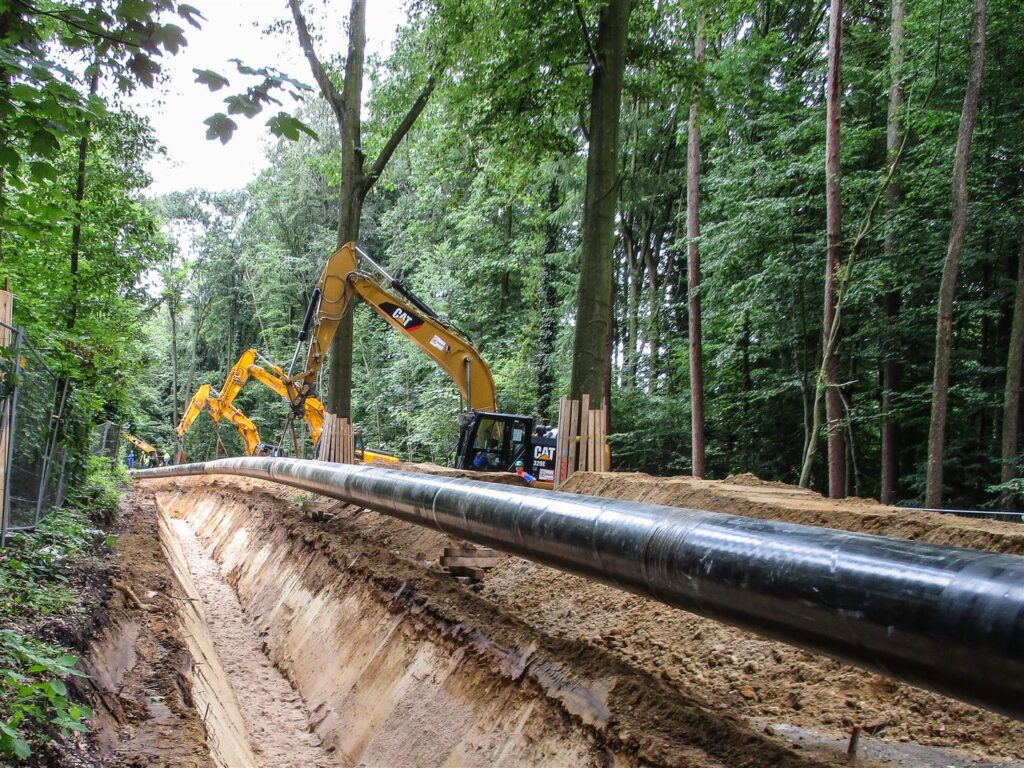 Eine Pipeline wird durch einen Waldabschnitt verlegt. Mehrere gelbe Bagger arbeiten daran, die Rohre an ihre Position zu bringen.