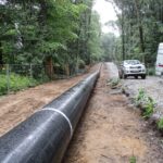 Eine Pipeline, die entlang einer Schotterstraße im Wald verläuft. Ein Bauwagen und ein Fahrzeug stehen daneben. Der Rohrstrang ist lang und scheint für den Einbau vorbereitet zu werden.