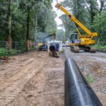 Eine Baustelle im Wald, auf der ein gelber Kran ein Rohrstück hebt. Arbeiter in blauen Anzügen sind bei der Installation der Pipeline beteiligt.