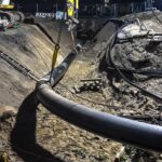 Nachtaufnahme einer Pipeline-Baustelle mit einem gelben Bagger, der eine Rohrleitung in einen Graben hebt. Baustellenbeleuchtung sorgt für Sichtbarkeit.