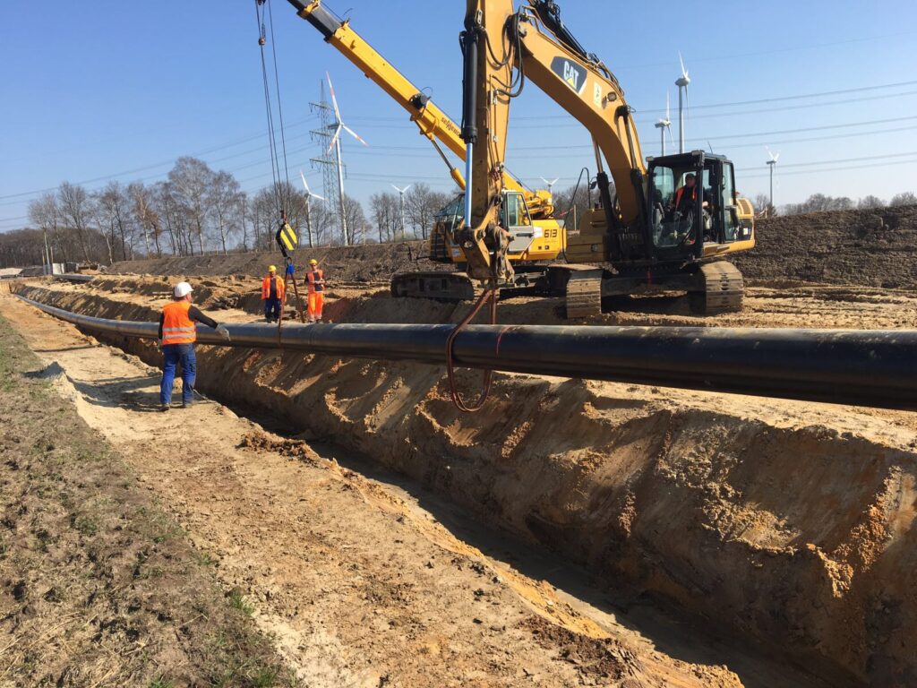 Eine Baustelle in einer offenen Landschaft mit mehreren großen Rohrabschnitten, die verlegt werden. Ein Arbeiter in blauer Schutzkleidung überwacht das Heben eines Rohrs durch einen großen gelben Bagger mit Greifvorrichtung. Im Hintergrund sind Windkraftanlagen zu sehen.