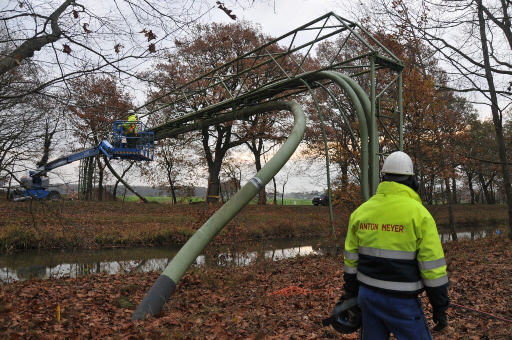 Eine über dem Boden schwebende Rohrbrücke ist teilweise beschädigt, einige Rohre sind verbogen und hängen lose herab. Arbeiter in einem Hebebühnenfahrzeug inspizieren die Struktur, während ein Mann in einer gelben Schutzjacke mit der Aufschrift „Anton Meyer“ die Szene beobachtet.