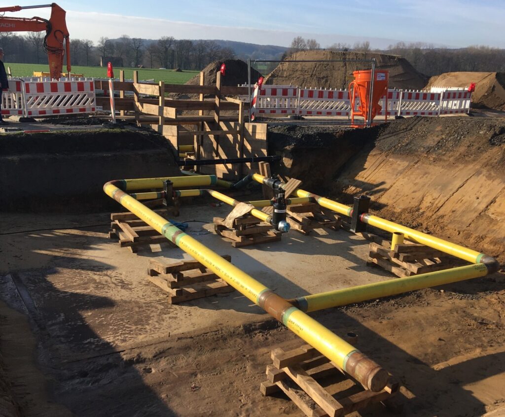 Eine Baustelle mit mehreren gelben Rohren, die durch schwarze Verbindungselemente ergänzt werden. Ein Arbeiter ist mit Schweißarbeiten beschäftigt, während im Hintergrund eine mobile Druckluftanlage und Baumaterialien bereitstehen.