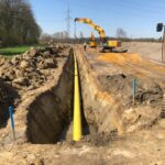 Eine Baustelle in einer ländlichen Umgebung mit einer tiefen Baugrube. Eine gelbe Versorgungsleitung wird in den Graben gelegt. Ein Bagger hebt Erdmaterial aus und bewegt sich entlang der Baustelle.