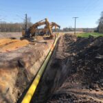 Eine Pipeline-Baustelle neben einer Eisenbahnlinie. Große Bagger arbeiten daran, eine lange Pipeline entlang einer Grube zu verlegen. Oberleitungen und Masten deuten darauf hin, dass die Baustelle in der Nähe eines elektrifizierten Schienennetzes liegt.