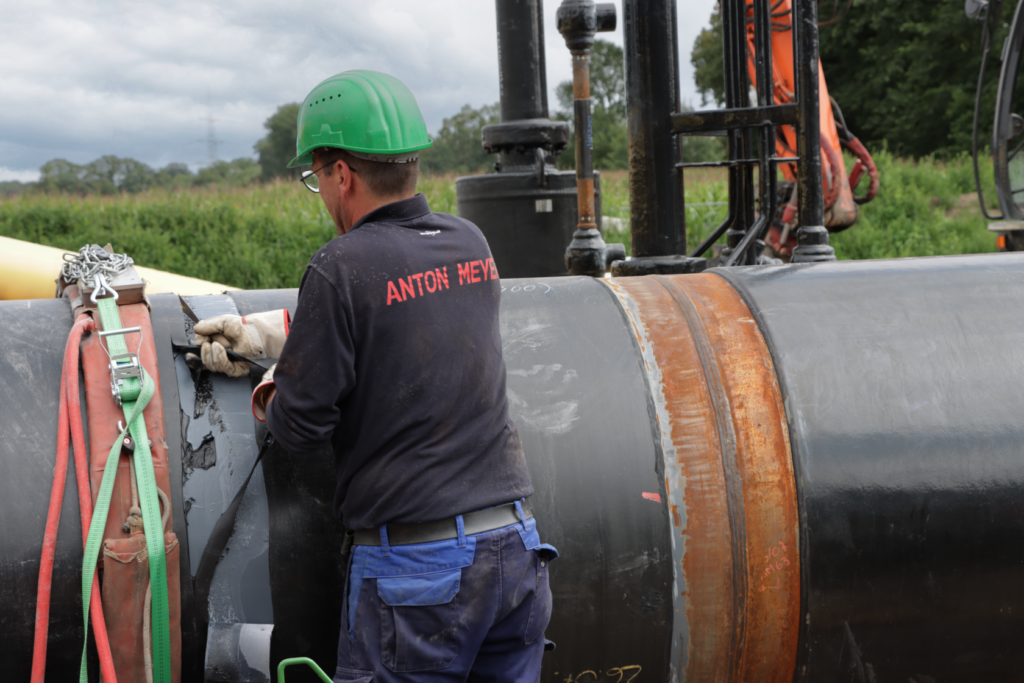 Ein Arbeiter mit grünem Schutzhelm und dunkler Kleidung mit der Aufschrift ANTON MEYER bringt im Freien, umgeben von grünem Gras und Industrieanlagen, Material an einer großen Rohrleitung an.
