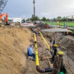 Eine Pipeline mit gelben Enden wird in einer offenen Baugrube verlegt, Arbeiter sind mit Schweißarbeiten beschäftigt.