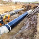 Eine große schwarze Pipeline wird in einem tiefen Graben mit Hilfe eines Minibaggers verlegt. Treppen und Geländer bieten Zugang zur Baustelle.