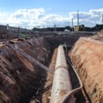 Eine lange Pipeline, bereits im Graben verlegt, mit angrenzenden Bauzäunen und einer Eisenbahnstrecke im Hintergrund.