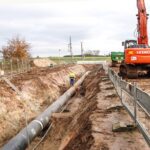 Ein Hitachi-Bagger mit orangem Gehäuse steht auf einer Baustelle neben einem tiefen Graben mit verlegter Pipeline, während ein Bauarbeiter den Fortschritt überprüft.