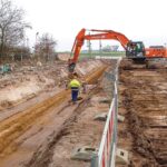 Ein Arbeiter mit Schutzkleidung steht in einem fast fertiggestellten Pipeline-Graben, während ein Hitachi-Bagger Erdreich bewegt.