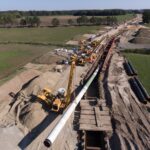 Ein langes Pipeline-Segment wird von mehreren gelben Kranen in einen Graben abgesenkt, mit gestapelten Rohren auf der Seite und einer weitläufigen Baustelle im Hintergrund.