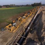 Eine Baustelle mit mehreren gelben Rohrverlegefahrzeugen, die eine Pipeline in einen vorbereiteten Graben heben. Der Blick geht entlang einer weiten Landschaft mit grünen Feldern.