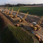 Eine Luftaufnahme zeigt eine lange Baustelle mit mehreren Rohrverlegefahrzeugen, die eine Pipeline verlegen. Der Hintergrund zeigt eine ländliche Landschaft mit Windrädern.