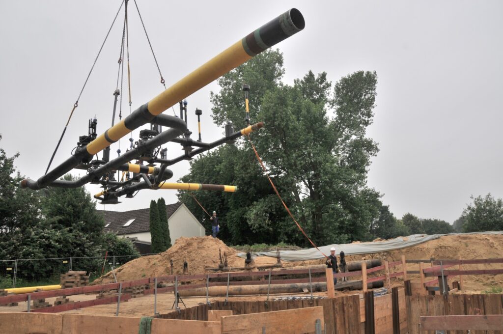 Eine große Rohrverbindung mit mehreren Ventilen wird von einem Kran in eine vorbereitete Baugrube abgelassen. Arbeiter sichern den Ladevorgang mit langen Führungsseilen. Der Himmel ist bewölkt, und im Hintergrund sind Wohnhäuser zu sehen.