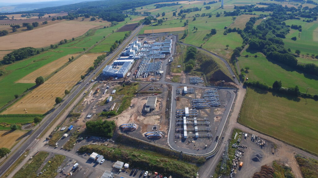 Luftaufnahme einer großen Baustelle, umgeben von grünen Feldern und Ackerland, mit mehreren Gebäuden, Baumaterialien und Fahrzeugen, die in der Gegend verstreut sind. Straßen und Bäume säumen die Baustelle.