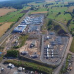 Luftaufnahme einer großen Baustelle, umgeben von grünen Feldern und Ackerland, mit mehreren Gebäuden, Baumaterialien und Fahrzeugen, die in der Gegend verstreut sind. Straßen und Bäume säumen die Baustelle.