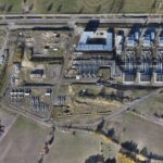 Luftaufnahme einer großen Industrieanlage mit mehreren rechteckigen Strukturen, Rohren und Maschinen, umgeben von Feldern, Straßen und etwas Vegetation. Am Boden sind die Schatten von Gebäuden und Bäumen zu erkennen.