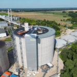 Aerial view of a large cylindrical industrial storage tank with a crane next to it, surrounded by smaller tanks, buildings and green fields in a rural area.
