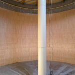 Ein großer, leerer, runder Innenraum mit glatten Holzwänden, einem grauen Betonboden und einer hohen weißen Säule in der Mitte, die bis zur Decke reicht.