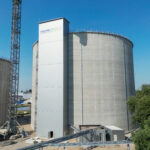 Ein großes zylindrisches Betonsilo steht neben einem hohen rechteckigen Gebäude mit dem Logo der Eraslan-Gruppe. In der Nähe sind Baugeräte und Container unter einem strahlend blauen Himmel zu sehen.